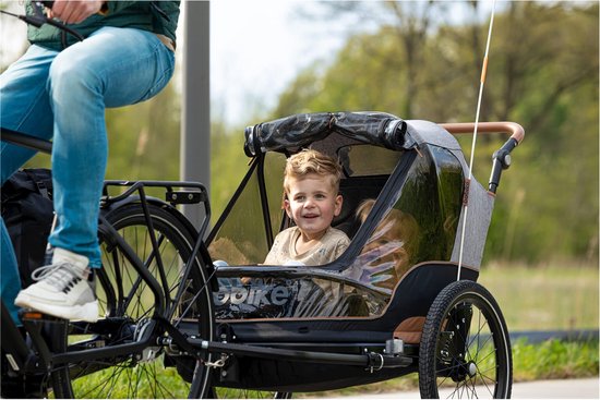 Een jongetje kijkt lachend uit een fietskar die achter een fiets hangt, met een persoon op de fiets.