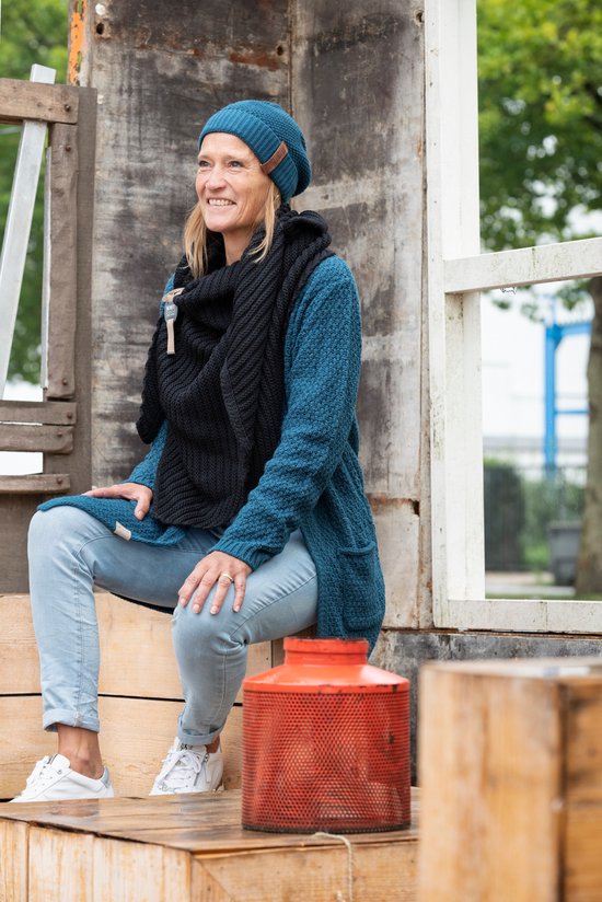 Vrouw met een zwarte gebreide sjaal, petrolblauwe vest en bijpassende muts. Ze draagt een spijkerbroek.