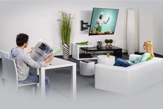 Een zwenkbare tv-muurbeugel gemonteerd aan de muur, met een tv erop. Een man leest aan een tafel, een vrouw zit op de bank.