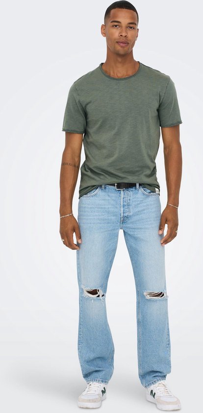 Man met tatoeage op arm, gekleed in groen T-shirt, zwarte riem en lichtblauwe jeans met gaten.