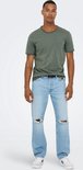 Man met tatoeage op arm, gekleed in groen T-shirt, zwarte riem en lichtblauwe jeans met gaten.