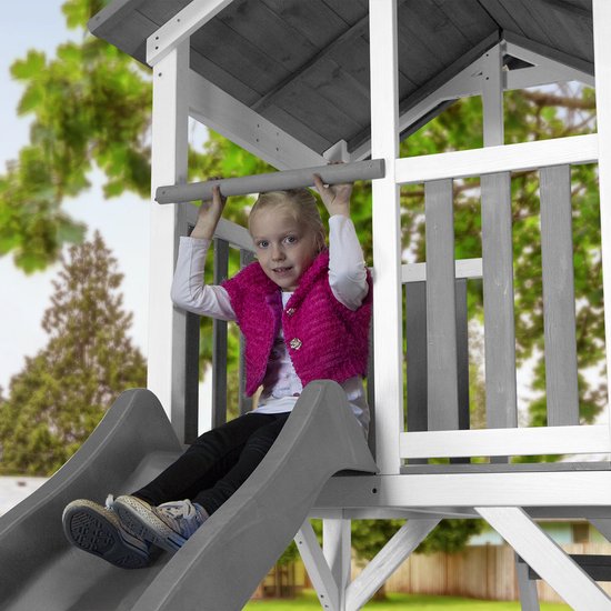 Meisje met felroze vest hangt aan een paal boven een grijze glijbaan in een speelhuis.
