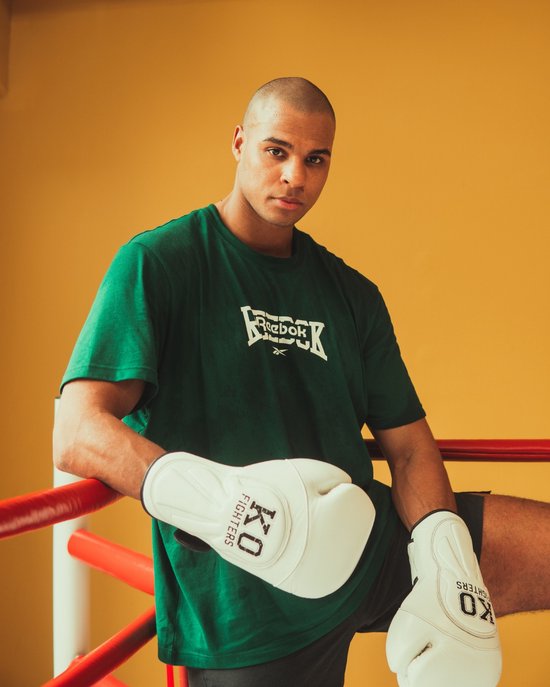 Een man met een groene t-shirt en witte KO FIGHTERS bokshandschoenen in een bokskooi.