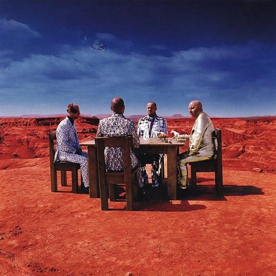 Vier mannen in opvallende pakken zitten aan een tafel in een woestijnlandschap onder een blauwe lucht. De pakken zijn bedrukt met patronen.