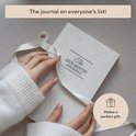 Linnenkleurige 'The Five Minute Journal' met witte linten, wordt vastgehouden door een persoon.