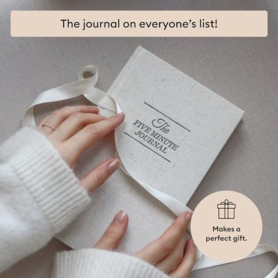 Linnenkleurige 'The Five Minute Journal' met witte linten, wordt vastgehouden door een persoon.