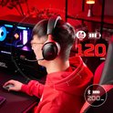 Man met rode hoodie draagt een zwarte en rode koptelefoon terwijl hij een pc-game speelt, met batterij- en connectiviteitsindicatoren.