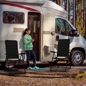 Vrouw leunt tegen camper bij twee zwarte campingstoelen.