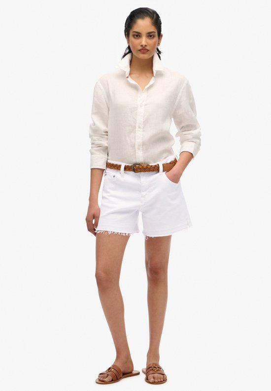 Model in wit denim short met rafelrand, witte blouse en bruine riem.