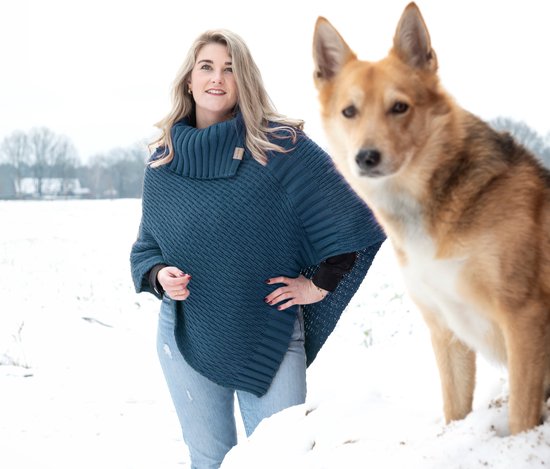Blauwe gebreide poncho met hoge kraag, gedragen door een vrouw met blond haar in de sneeuw, naast een hond.