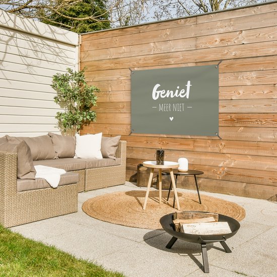Donkergroene banner met tekst 'Geniet - Meer Niet -' hangend aan een houten schutting in een tuin met loungeset.