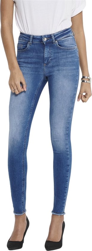 Vrouw met lang bruin haar, draagt blauwe skinny jeans met rafelige zoom en witte top, gedragen met zwarte pumps.