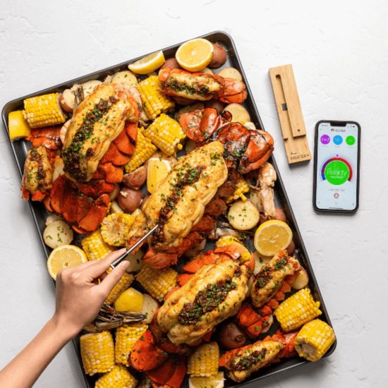 Gegrilde kreeftstaarten en maïskolven op een bakplaat, met citroenpartjes en een Meater thermometer en app.