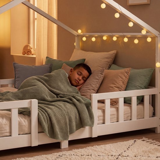 Enfant dormant dans un lit maison blanc avec barreaux, décoré de guirlandes lumineuses.