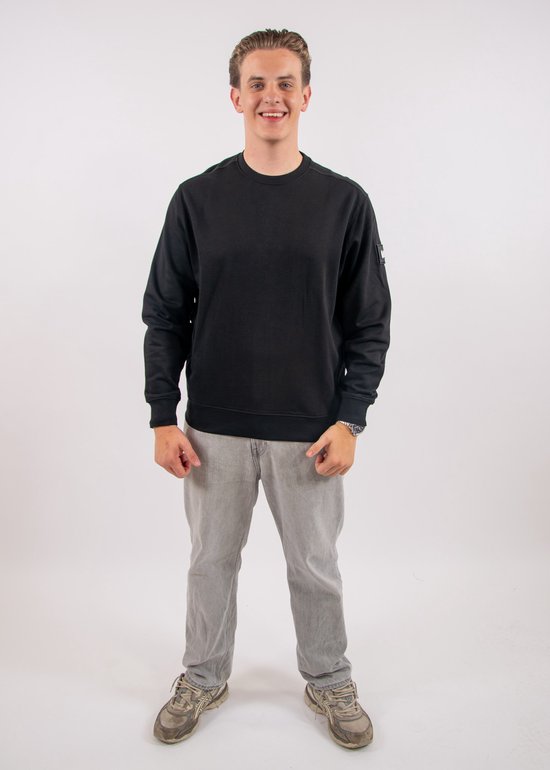 Model ten voeten uit in een zwarte sweater met ronde hals, grijze broek en sneakers.