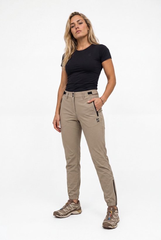 Femme bras croisés portant un pantalon outdoor beige avec fermetures éclair noires et logo sur le côté.