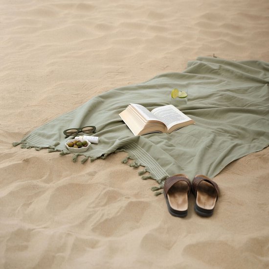 Groene hamamdoek uitgespreid op het strand met een boek, zonnebril, olijven en slippers.