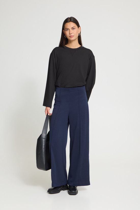 Model in een zwarte top en donkerblauwe broek met wijde pijpen, staand met een zwarte tas.