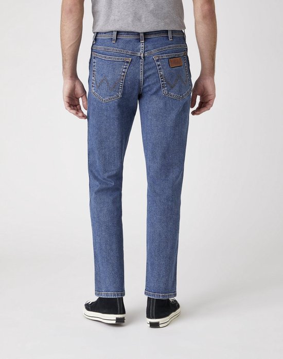Achterkant van een man in een blauwe Wrangler jeans, met zwarte Converse sneakers.