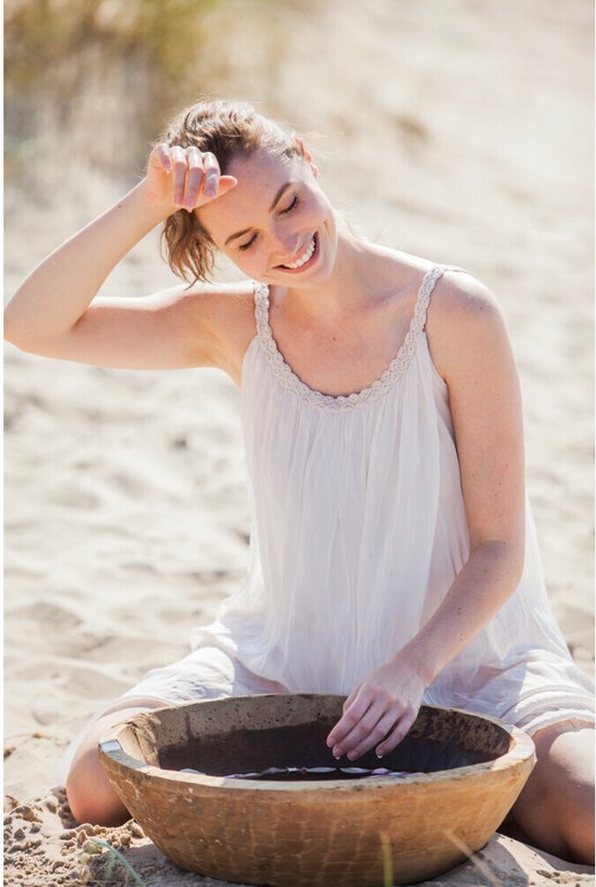 Vrouw met bruin haar, lachend, met witte jurk, op het strand, met hand in houten kom.