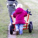 Une brouette rouge à quatre roues pleines jaunes est tirée par deux enfants.