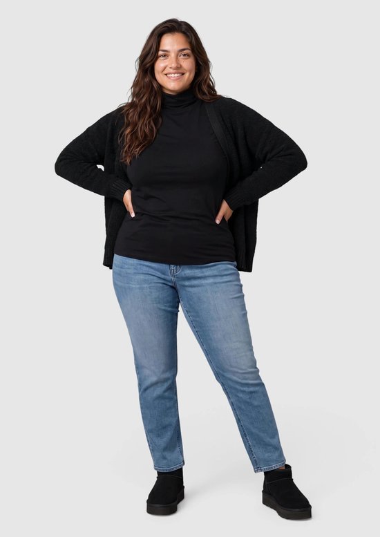 Vrouw draagt een zwarte coltrui onder een bijpassend zwart vest, blauwe jeans en zwarte instappers.