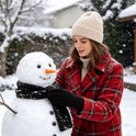 Vrouw in rood geruite jas en beige muts draagt zwarte handschoenen en versiert een sneeuwpop.