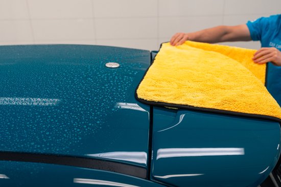 Een persoon die een felgeleende microvezel droogdoek vouwt op de teal blauwe auto, met waterdruppels.