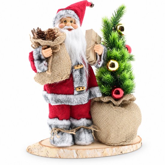 Figurine du Père Noël en tenue rouge à bordure grise en fourrure, tenant un sac de pommes de pin et un cadeau. À côté de lui, un sapin de Noël illuminé dans un sac en jute.