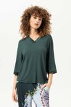 Model in een donkergroene top met driekwart mouwen en een V-hals versierd met satijn. De top is gecombineerd met een broek met een kleurrijke, botanische print.