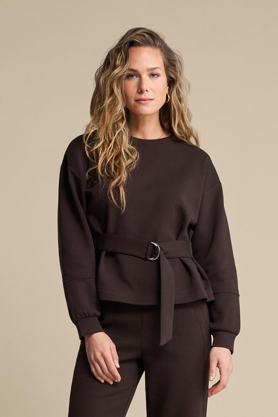 Model met lang blond golvend haar draagt een donkerbruine sweater met ceintuur en wijde broek.