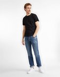 Man met bruin haar, gekleed in een zwart T-shirt en een lichtblauwe spijkerbroek, staat met handen in zakken.