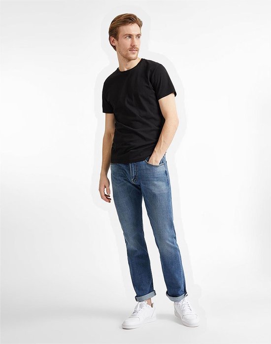 Man met bruin haar, gekleed in een zwart T-shirt en een lichtblauwe spijkerbroek, staat met handen in zakken.