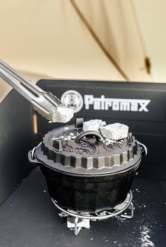 Tangen leggen witte briketten op de deksel van een zwarte Petromax Dutch oven. Buiten, met tentdoek op achtergrond.