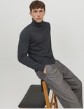 Man zittend op een stoel, draagt een donkergrijze coltrui en een grijze pantalon met krijtstrepen.