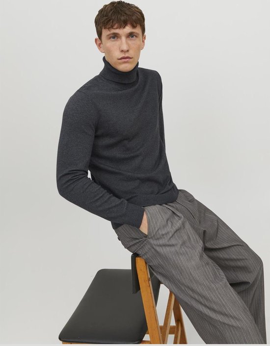 Man zittend op een stoel, draagt een donkergrijze coltrui en een grijze pantalon met krijtstrepen.
