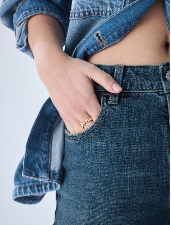 Model draagt een spijkerbroek met een denim top, de hand is in de broekzak met een gouden ring.
