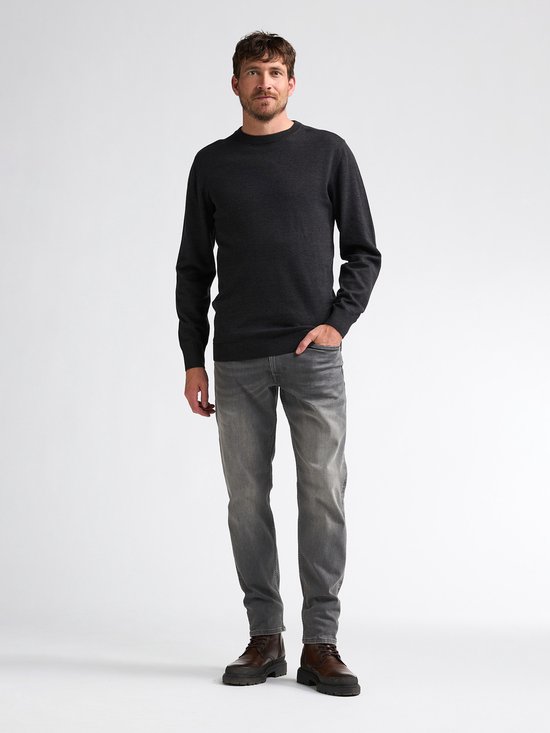 Man staat met zijn handen in de zakken van zijn grijze jeans, draagt een donkergrijze gebreide trui.