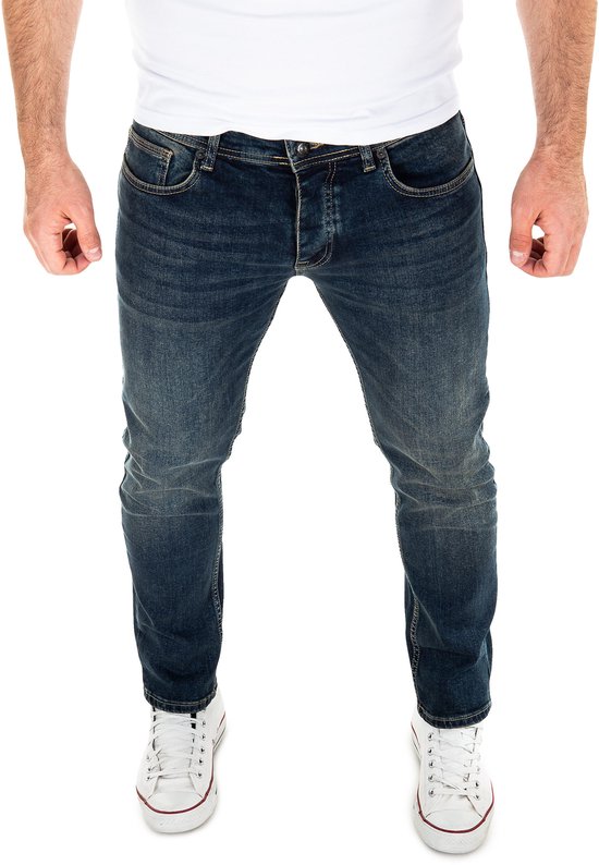Man met donkerblauwe jeans met stretch, 5 zakken, en witte sneakers.