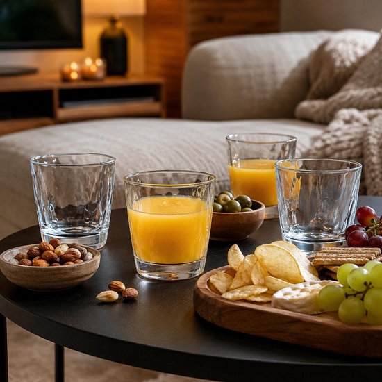 Quatre verres à motif ondulé, remplis de jus ou vides. Sur une table basse avec snacks et fruits.