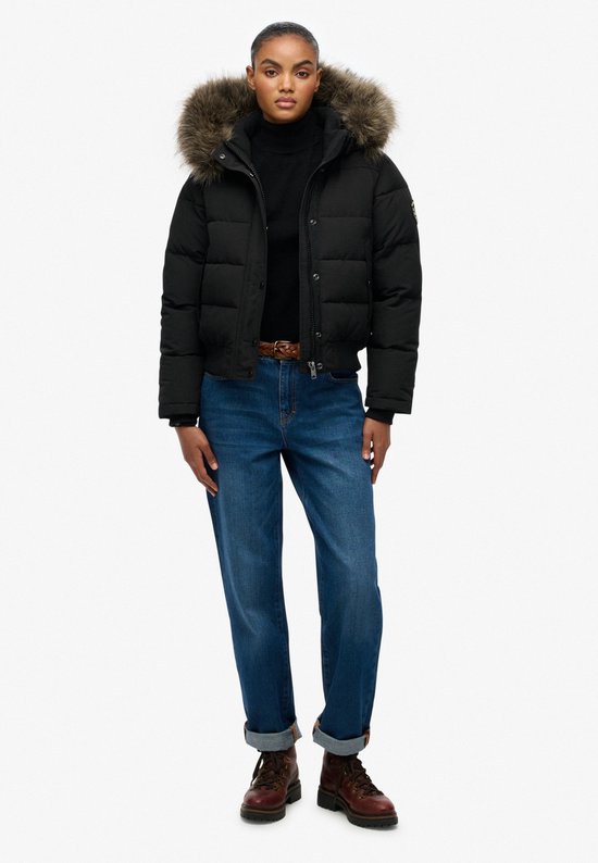 Model in een zwarte korte gewatteerde jas met bontkraag, zwarte coltrui, spijkerbroek en bruine veterlaarzen.