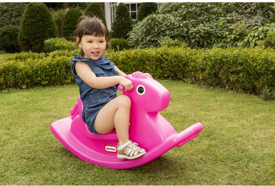 Een jong kind in een denim jurk op een roze Little Tikes hobbelpaard in een tuin.