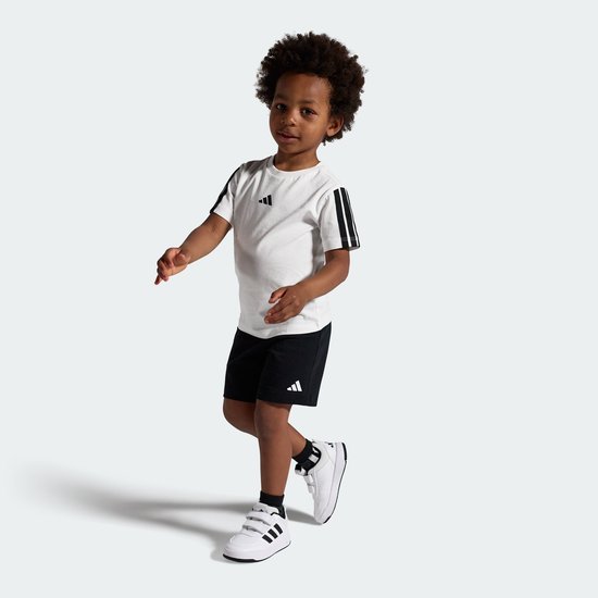 Een jongetje met krullend haar draagt een wit T-shirt met zwarte strepen op de mouwen en zwarte shorts.