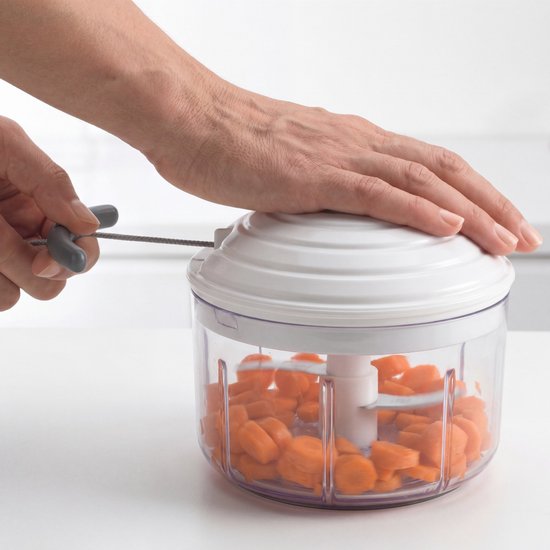 Een hand trekt aan de grijze hendel van de witte keukenmachine, gevuld met blokjes wortel.