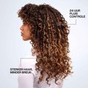 Profil d'une femme aux cheveux bruns ondulés avec reflets. Le texte indique 24h de contrôle des frisottis et des cheveux plus forts.