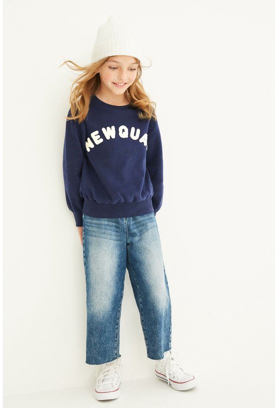 Model draagt een donkerblauwe trui met 'NEWQUA 1982' en een blauwe mom-jeans.