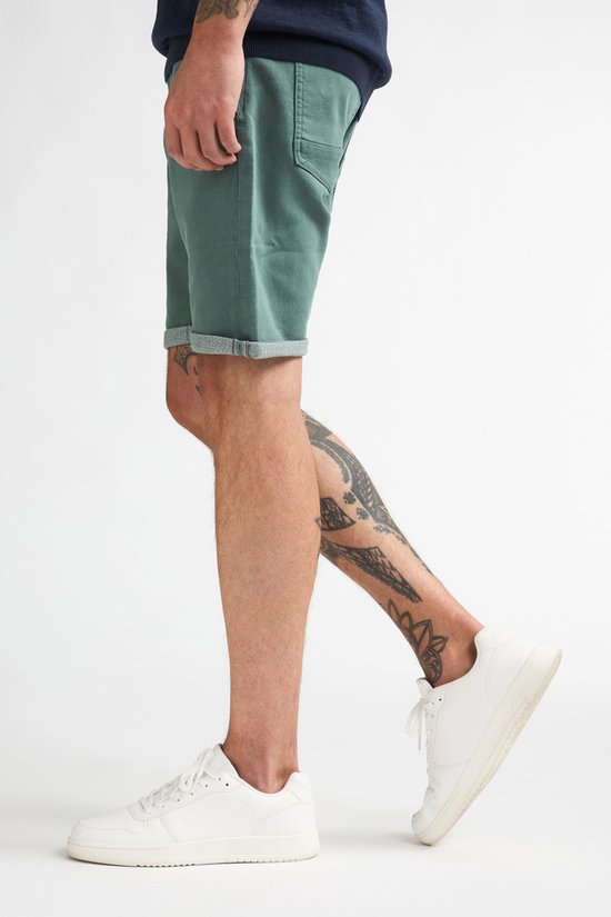 Zijaanzicht van groene slim fit stretch denim shorts met opgerolde zomen, gedragen door een model met tattoos.