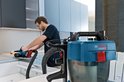 Blauwe Bosch GAS 18V-10 L stofzuiger op de voorgrond, terwijl een vakman op de achtergrond zaagt.