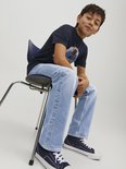 Jongen in donkerblauw T-shirt met print, zittend op stoel in lichtblauwe jeans. Draagt sneakers.