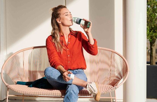 Vrouw in oranje blouse drinkt uit een Mepal waterfles met groene band.